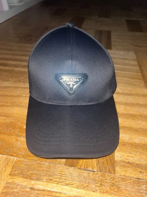 Svart keps från Prada - Snygg svart keps med det klassiska trekantiga metallmärket framtill. Kepsen har böjd skärm och justerbar rem bak. Tillverkad i ett glansigt, slitstarkt material som ger en exklusiv känsla. Perfekt för dig som vill ha en stilren accessoar med lyxig touch.