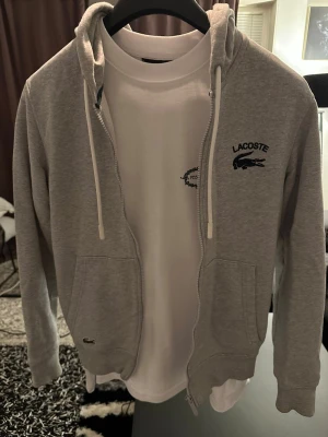 Grå zip-hoodie från Lacoste - Tvättat ett par gånger, köpte för 1657. Snygg grå zip-hoodie från Lacoste med huva och vita dragsnören. Tröjan har två fickor framtill och klassisk Lacoste-logga med krokodil på bröstet. Perfekt för en avslappnad och sportig stil. Tillverkad i mjukt material för extra komfort.