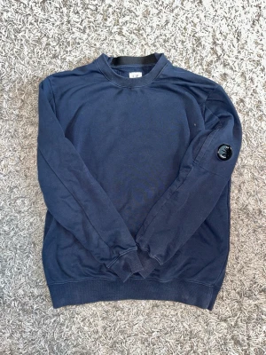 Cp company sweatshirt - Snygg mörkblå sweatshirt från C.P. Company med rund halsringning och ribbade muddar. Tröjan har en ikonisk linsdetalj på ena ärmen och är tillverkad i mjukt bomullsmaterial. Perfekt för en avslappnad och stilren look.