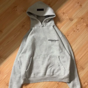 Grå Essentials Set - Snygg ljusgrå hoodie från Essentials Fear of God. Tröjan har svart logotyptryck på bröstet och en relaxed passform. Mjuk o skön bomull perfekt för chill dagar.