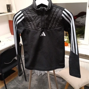 Svart Adidas half zip tröja 140 - Svart långärmad tröja från Adidas med quiltad överdel och klassiska vita ränder på ärmarna. Half zip framtill och Aeroready-tryck nedtill. Perfekt för träning eller chill, med sportig look och bekväm passform.
