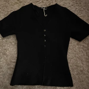 Svart v-ringad topp från Gina Tricot - Svart v-ringad topp från Gina Tricot med korta ärmar och snygga knappar längs hela framsidan. Toppens passform är figurnära och den är tillverkad i ett mjukt material som känns skönt mot huden. Perfekt att styla med jeans eller kjol för en clean look.