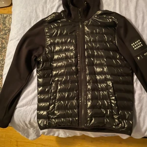 Svart pufferjacka Peak Performance - Svart pufferjacka från Peak Performance med glansig, quiltad framsida och mjuka ärmar. Jackan har huva, dragkedja framtill och två fickor. Snygg logga på ärmen och modern, sportig look. Perfekt för dig som gillar stilren och funktionell design.