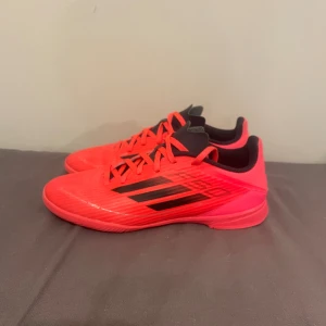 Adidas F50 röda inne fotbollsskor - Säljer ett par Adidas F50 fotbollsskor i en riktigt snygg röd färg med svarta detaljer och klassiska tre ränder på sidan. Skorna har snörning, låg profil och är tillverkade i syntetmaterial för bästa känsla på planen. Perfekta för dig som vill sticka ut på matchen!