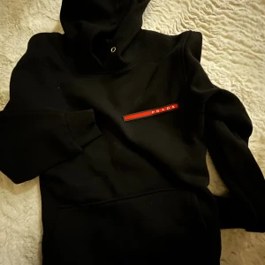 Svart hoodie från Prada - Säljer en svart hoodie från Prada med ikonisk röd logga på bröstet. Tröjan har huva med snörhål och en stor magficka. Perfekt för dig som vill ha en clean och lyxig streetwear-look. Storlek S-M
