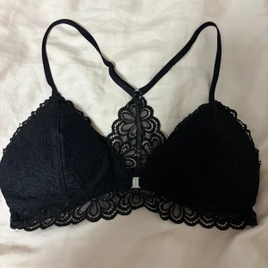 Svart spets bralette från Hollister - Supersnygg svart bralette från Hollister, modell Gilly Hicks. Den har tunna axelband och är täckt av blommig spets med fina detaljer längs kanterna. Perfekt för att ge en extra touch till din outfit. Materialet är mjukt och stretchigt för skön passform.