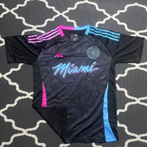 Inter Miami CF svart fotbollströja S - Svart Inter Miami CF fotbollströja från Adidas i slim fit. Tröjan har korta ärmar, rosa och blå detaljer på axlar och sidor, samt klubbmärke och Miami-tryck framtill. Tillverkad i lätt polyester som andas, perfekt för match eller träning.