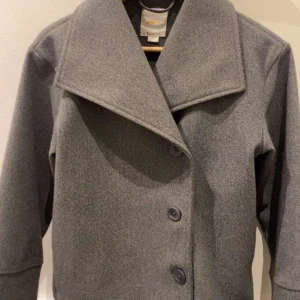 Grå kappa - Snygg grå kappa med bred krage och tre stora knappar framtill. Jackan har långärmade raka ärmar och är tillverkad i mjukt ullmaterial som håller dig varm under kyliga dagar. Perfekt för dig som gillar stilren och klassisk look.