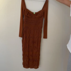 Brun långärmad smockad klänning - Säljer en snygg brun långklänning med smockad struktur och långärmade ärmar. Klänningen har en figurnära passform och ett rynkat bröstparti som ger en cool och trendig look. Perfekt för dig som gillar stilrena och unika detaljer.