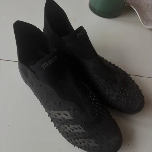 Adidas Predator svarta fotbollsskor - Säljer ett par svarta Adidas Predator fotbollsskor med coola gummitaggar på ovansidan för extra grepp på bollen. Skorna har en strumpliknande passform utan snörning och en slitstark sula med blå detaljer och dobbar för bra fäste på planen.
