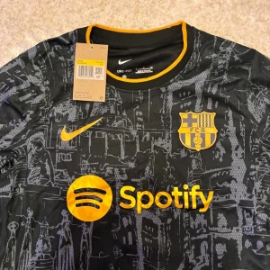 Barcelona svart matchtröja Nike S - Barcelona fotbollströja i svart med grått grafiskt mönster inspirerat av stadens arkitektur. Gula detaljer på krage, ärmslut, Nike-logga och sponsortryck. FC Barcelona-emblem på bröstet, Spotify-logga framtill och UNHCR-logga baktill. Tillverkad i lätt, ventilerande Dri-FIT-material.