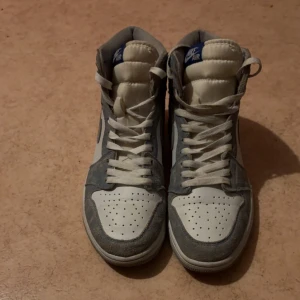 Nike Air Jordan 1 High OG vit/grå - Säljer ett par Nike Air Jordan 1 High OG i vitt och ljusgrått. Skorna har klassisk hög siluett, snörning och perforerad tå. Ovandelen är i skinn och mocka med grå Swoosh och Air Jordan-logga på sidan. Perfekta för dig som gillar streetwear och sneakerskultur.