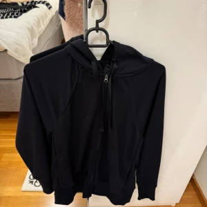 Svart Nike hoodie Dri-Fit XS - Svart hoodie från Nike i Dri-Fit material, perfekt för träning. Tröjan har huva med snörning, dragkedja framtill och långa ärmar. Snygg och sportig look som funkar till chill eller gym. Materialet är mjukt och andas bra.