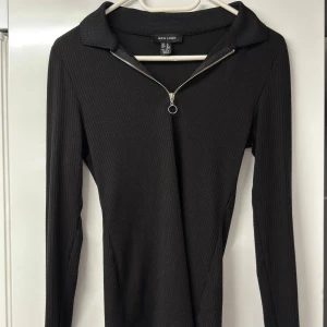 Svart ribbad långärmad polotopp från New Look - Svart ribbad polotopp från New Look i storlek M. Toppen har lång ärm, krage och en snygg silverfärgad half zip-dragkedja framtill med ringdetalj. Materialet är mjukt och stretchigt, perfekt för en clean och stilren look.