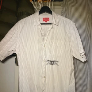 Randig Supreme x Anti skateboard Hero skjorta  - Supreme x Anti Hero kortärmad skjorta i vitt med tunna blå ränder. Tillverkad i 100% bomull för en skön känsla. Pris kan diskuteras #archive #grisch #supreme #antiskateboardheros #skjorta #archive #still #tröja #oldmoney #laidback 