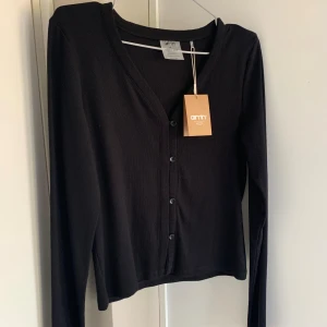 Svart kofta från Aim'n med knappar - Svart kofta från Aim'n med v-ringning och knäppning framtill. Långärmad och i mjukt, tunt material som känns skönt mot huden. Perfekt att slänga över en topp eller bära stängd för en clean look.