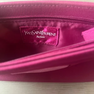 Rosa kuvertväska från Yves Saint Laurent - Snygg rosa kuvertväska från Yves Saint Laurent Parfums med hjärtformad detalj och YSL-logga på framsidan. Väskan är tillverkad i ett glansigt syntetmaterial och har dragkedja upptill. Perfekt för att bära dina essentials med stil. Kan användas som smink väska limit edition.