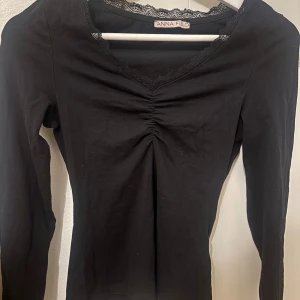 Svart långärmad topp från Anna Field - Svart långärmad topp från Anna Field med v-ringning och spetskant runt halsen. Toppen har rynkdetalj framtill som ger en snygg form. Perfekt att styla med jeans eller kjol 