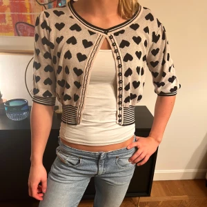 Beige kofta med svarta hjärtan - Supergullig croppad kofta i beige med svarta hjärtan över hela, samt svarta och vita ränder vid muddar och nederkant. Koftan har knappar framtill och trekvartsärmar. Perfekt för dig som gillar söta detaljer och vill sticka ut lite extra.