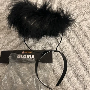 Svart gloria-diadem från Partyking - Säljer ett svart gloria-diadem från Partyking. Diademet har en fluffig gloria i svart fjäderliknande syntetmaterial som sitter på en tunn svart båge. Perfekt accessoar för temafest eller maskerad.