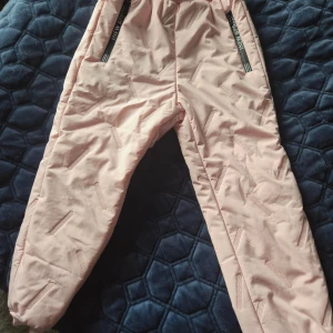 Ljusrosa täckbyxor med dragkedjor - Säljer ett par ljusrosa täckbyxor från New Sport, storlek 120. Byxorna har svarta dragkedjor med logga, elastisk midja och är fodrade med mjuk teddy på insidan. Materialet är 100% polyester och de har diskreta mönsterdetaljer på benen.