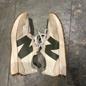 New Balance 327 - Snygga New Balance sneakers med beige och vita paneler, olivgrön logga på sidan och grov brun yttersula. Skorna har snörning, rund tå och är tillverkade i en mix av mocka och syntetmaterial. Perfekta för dig som gillar retrostil och streetwear.