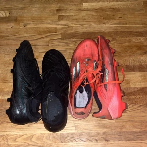 Nike och Adidas fotbollsskor - Två par fotbollsskor: ett par helsvarta Nike med snörning och rund tå, samt ett par röda Adidas med svarta och silvriga ränder. Båda har dobbar för gräsplan och är tillverkade i syntetmaterial. Perfekta för dig som vill sticka ut på planen.   Skriv vid fråga om pris 