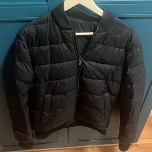 Svart pufferjacka från J.Lindeberg - Snygg svart pufferjacka från J.Lindeberg med diskret logga på bröstet. Jackan har quiltad design, dragkedja framtill och ribbade muddar vid ärmslut och midja. Perfekt för kyliga dagar och ger en clean, sportig look. Orginal pris 1499.