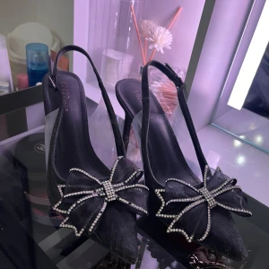 Svarta pumps med strassrosett - Supersnygga svarta pumps från INN99 med spetsig tå och öppen häl. Skorna har en stor rosett framtill dekorerad med glittrande strass, vilket ger en extra glamorös touch. Perfekta för dig som vill sticka ut med elegans och stil.