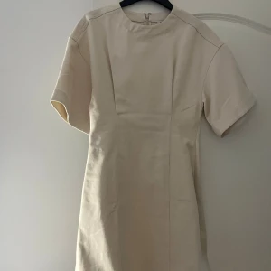 Beige kortklänning från & Other Stories - Snygg beige kortklänning från & Other Stories med rund hals och korta ärmar. Klänningen har en stilren, rak passform och är tillverkad i bomull med diskret struktur. Dragkedja bak för enkel påklädning. Perfekt för dig som gillar minimalistisk stil.