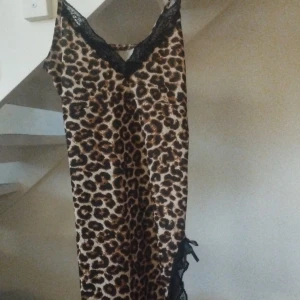 Leopardmönstrad långklänning med spets - Snygg långklänning med leopardmönster i bruna och svarta toner. Klänningen har tunna axelband och v-ringad hals med svart spetsdetalj. Nedtill finns en slits med svart spets och rosett. Materialet är glansigt och känns lyxigt.