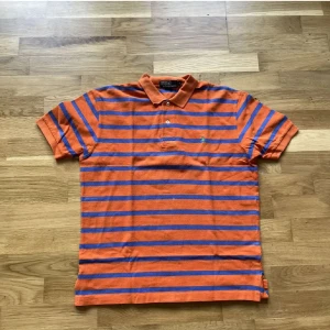 Orange och blå randig pikétröja Polo Ralph Lauren - Orange pikétröja från Polo Ralph Lauren med blå horisontella ränder och klassisk krage. Vissa defekter som visas på bilderna 