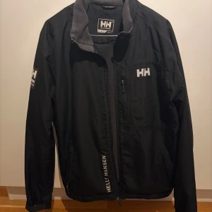 Svart vindjacka Helly Hansen M - Svart vindjacka från Helly Hansen i storlek M. Jackan har fleecefodrad krage, dragkedja framtill, justerbara ärmslut med kardborre och flera loggor på bröst, ärm och rygg. Perfekt för dig som gillar sportig och clean stil.    (Ända felet med den är visat på sista bilden när ”plasten” på kardborren, men detta märks inte🤙)
