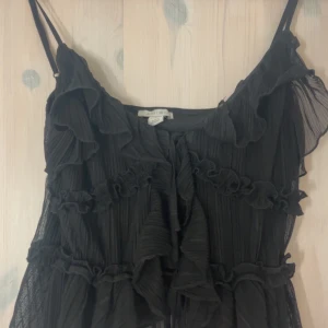 Svart volangklänning från Amisu/new yorker - Supersöt svart klänning från Amisu med tunna axelband och flera lager volanger. Klänningen har en luftig känsla med plisserat tyg och romantiska detaljer framtill. Perfekt för dig som gillar en feminin och trendig stil.