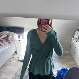 Grön omlottblus med v-ringning - Säljer en snygg grön blus med omlott-design och v-ringning. Toppen har långa ärmar och ett smickrande resår i midjan som ger en fin siluett. Materialet känns mjukt och lätt, perfekt för dig som gillar stilrena och bekväma plagg.