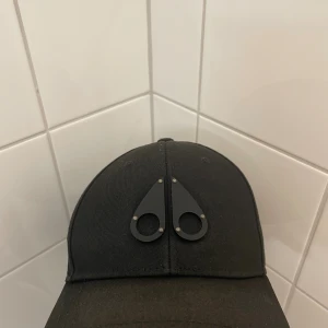 Svart keps från MOOSE KNUCKLES - Svart keps från Moose Knuckles med deras ikoniska metall-logga framtill. Justerbar rem baktill med logodetalj. Klassisk böjd skärm och stilren design, perfekt för dig som gillar snygga accessoarer.