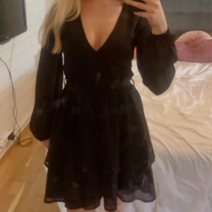 Svart omlottklänning med v-ringning - Snygg svart kortklänning med omlott och djup v-ringning. Klänningen har långa, lätt transparenta ärmar och markerad midja med knytband. Kjolen är volangig och ger en flowy känsla. Perfekt för dig som gillar en stilren och trendig look.
