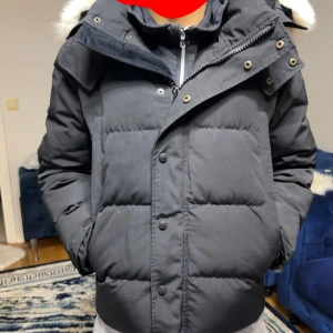 Svart Canada Goose - Svart pufferjacka från Canada Goose med dunfyllning och fluffig päls på huvan. Jackan har dragkedja, knappar och logotyp på ärmen. Snygg, varm och perfekt för kalla dagar. Modellen är normal i passningen och har långa ärmar.Har även påsen till den dm för mer bilder 