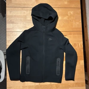 Svart Nike tech hoodie  med dragkedja - Svart hoodie från Nike med hel dragkedja framtill och två fickor med dragkedja. Tröjan har huva, lång ärm och diskret Nike-logga på bröstet. Perfekt för chill eller träning, tillverkad i mjukt och stretchigt material.