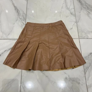 Plisserad brun skinnkjol FB Sister XS - Snygg plisserad kortkjol från FB Sister i en varm brun färg. Kjolen är gjord i skinnimitation med mjukt beige foder på insidan. Perfekt för dig som gillar en trendig och edgy look med lite extra attityd.