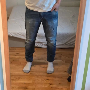 Blå jeans med slitningar från Diesel - Säljer ett par blå jeans med snygga slitningar och tvättade detaljer på låren och knäna. Modellen har klassisk femficksdesign, normal passform och raka ben. Jeansen är tillverkade i jeansmaterial och har en cool, avslappnad vibe.