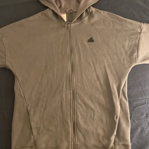 Grå hoodie från Adidas XL I BARN STORLEK - Säljer en grå hoodie från Adidas i storlek XL för tonåringar. Tröjan har hel dragkedja framtill, huva och ett diskret Adidas-logga på bröstet. Perfekt för chill dagar eller när du vill ha en enkel och stilren look. Aldrig använd.