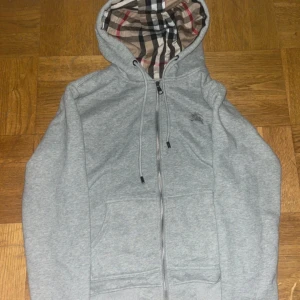 Burberry kofta - Grå hoodie inspirerad av Burberry med dragkedja och klassisk huva. Insidan av luvan har det ikoniska rutmönstret i beige, svart, vitt och rött. Tröjan har snörning i huvan, två fickor fram och ribbade muddar. Perfekt för en chill och stilren look. Tröjan har storlek L men sitter som storlek S och passar också till storlek M. 100% Bomull.