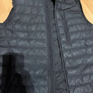 Svart dunväst från Peak Performance - Svart quiltad dunväst från Peak Performance med diskret logga på bröstet och dragkedja framtill. Västen har en bröstficka med dragkedja och är tillverkad i lätt polyester. Perfekt för lager-på-lager under kyliga dagar. liten defekt se bild ovan 
