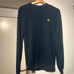 Marinblå sweatshirt från Lyle & Scott - Snygg marinblå sweatshirt från Lyle & Scott med klassisk gul logga på bröstet. Tröjan har rund hals, långa ärmar och ribbade muddar. Perfekt för dig som gillar stilrena och enkla plagg med en touch av sportig vibe.