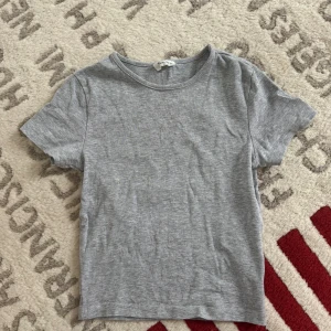 Grå basic t-shirt från Gina Tricot - Enkel och stilren grå t-shirt från Gina Tricot. Modellen är klassisk med rund halsringning och korta ärmar. Tillverkad i mjuk bomull som känns skön mot huden. Perfekt att matcha med jeans eller shorts för en clean look.