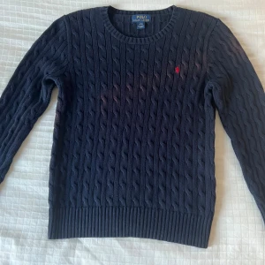 Mörkblå stickad tröja Polo Ralph Lauren - Snygg mörkblå stickad tröja från Polo Ralph Lauren med klassiskt kabelstickat mönster och röd broderad logga på bröstet. Rund halsringning och ribbade muddar vid ärmslut och nederkant. Perfekt för dig som gillar preppy stil.