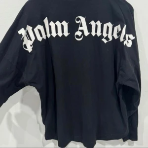 Svart långärmad tröja Palm Angels - Svart långärmad tröja från Palm Angels med stor vit logga längs ryggen och mindre logga tryckt vid halsen. Tröjan har rund halsringning och är gjord i mjuk bomull. Perfekt för dig som gillar streetwear och vill sticka ut med en ikonisk look.
