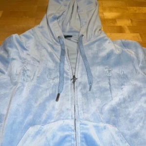 Ljusblå velour hoodie Juicy Couture - Supermjuk ljusblå hoodie från Juicy Couture i velour med dragkedja, snörning i huvan och broderad JC-logga på bröstet. Perfekt för en chill och trendig look.