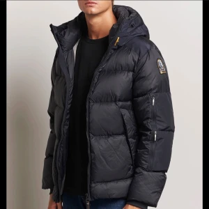 Svart dunjacka från Parajumpers - Snygg svart dunjacka från Parajumpers med huva och dragkedja. Jackan har en quiltad design, flera fickor med dragkedja och en patch på ärmen. Perfekt för kalla dagar och riktigt stilren look.